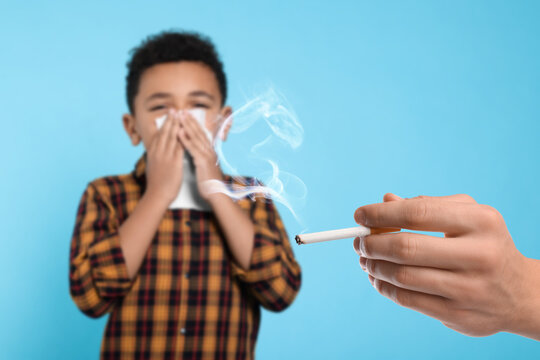 Passive smoking. Little boy covering his nose with tissue because of man smoking nearby on light blue background. Selective focus