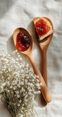 Two wooden spoons hold vibrant red jam, one atop a toasted bread slice; small white flowers rest nearby on a light fabric backdrop