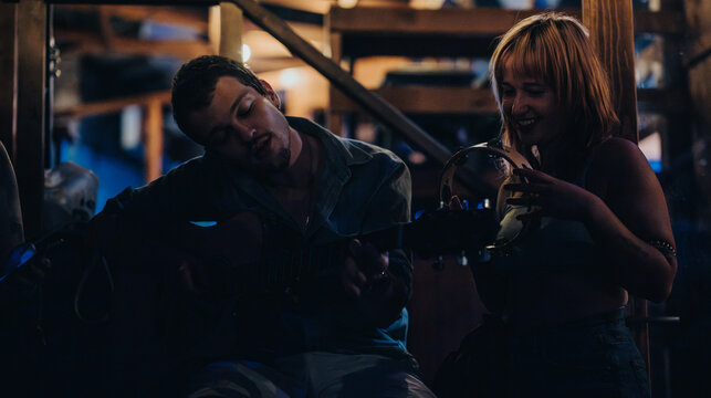 Two young adults immersed in a cozy musical jam session, strumming guitar and playing tambourine in a dimly lit, rustic environment that radiates joy and connection. - Powered by Adobe