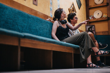 A pair of young friends sit comfortably on a bench in a trendy cafe, engaging in lively conversation and pointing at decorations. The atmosphere is casual and inviting.