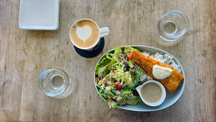 Enjoying crispy English-style fish and chips with tartar sauce at a café, meal set with a hot latte for lunch or afternoon tea — natural light, wooden table, glass water cup