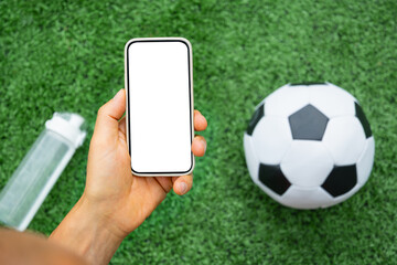 Man hand holds mobile phone with white blank screen on soccer field with ball on green grass. Modern technology with traditional sports. View from above. Empty display. Copy space.