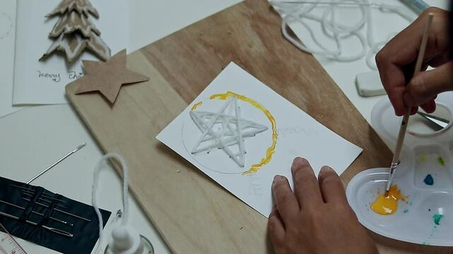 Hand painting yellow color in a circle around a sewn star shape yarn to create a parol design or a traditional Christmas lantern of the Philippines, handmade holidays greetings card