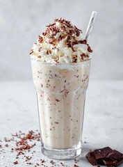 Creamy milkshake in a tall glass, topped with whipped cream and chocolate shavings, rests on a mottled grey surface with scattered chocolate pieces