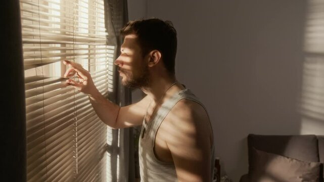 Side view shot of depressed army veteran looking through blinds in window with caring young woman embracing hm from back, scene lit by moody sunlight rays