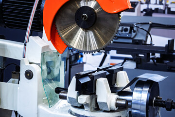 Close-up of a metal cutting saw with a circular blade and orange guard, showing the vise mechanism holding material. Industrial workshop equipment for precision cutting.