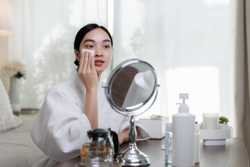 Young asian woman removing makeup with cotton pad in front of mirror