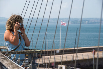 A female Street photographer holding a retro camera to her eye. Steel cables fan across the frame...
