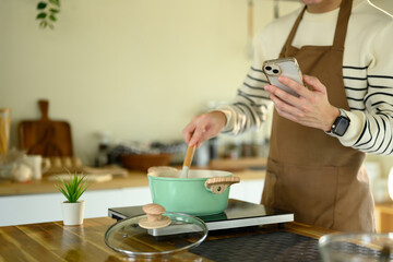 Young man stirs ingredients in a pot while referencing a recipe on his phone in a modern kitchen