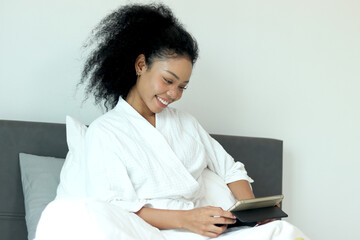 Beautiful woman with curly hair in white bathrobe using digital tablet after waking up in morning....