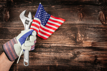 A workers gloved hand proudly holds tools and the American flag against a rustic wood background,...