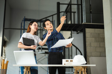 Asian man and woman architects collaborating with laptop and tablet while pointing at building structure on interior construction site.