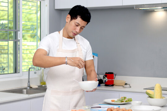 Happy Asian man with apron preparing breakfast meal in the kitchen. A man adding seasoning to the bowl for cooking tasty food meal at kitchen at home. Man cooking delicious food.