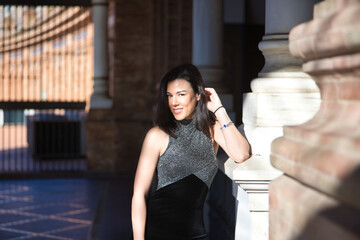 Portrait of young, beautiful, brunette woman, wearing a fancy black dress, looking at the camera, relaxed and calm, leaning on a marble column, receiving the sun's rays. Concept beauty, femininity.