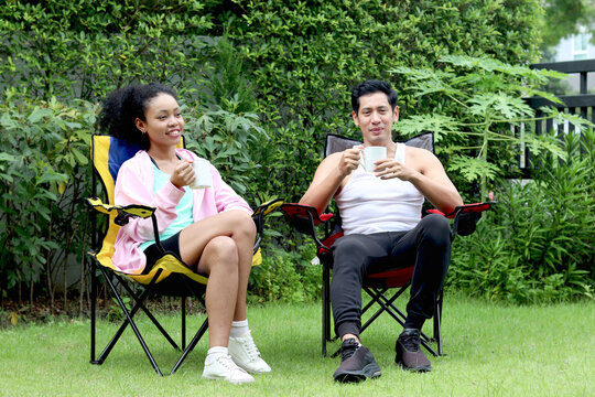 Happy couple camping in green park. Asian man with African woman with curly hair drinking coffee and spending time outdoor together in backyard. The lover sitting and talking. Diverse family.