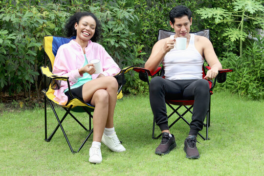 Happy couple camping in green park. Asian man with African woman with curly hair drinking coffee and spending time outdoor together in backyard. The lover sitting and talking. Diverse family. - Powered by Adobe