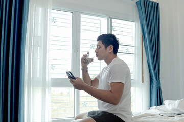 Asian man using smartphone while drinking almond milk, sitting in bed next to bedroom window. Man...