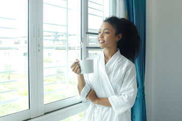 Beautiful curly hair woman in white bathrobe holding coffee mug while standing in the bedroom after...