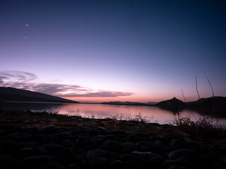Purple and pink gradient sky over tranquil water at twilight. Soft twilight glow reflects on the calm lake surface with dark foreground elements. Ideal for travel, nature, and website backgrounds.
