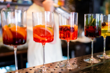 A row of five colorful cocktails in tall glasses sits on a bar counter, each garnished with fruits and herbs. The vibrant display, blurred background, and bright hues emphasize freshness atmosphere