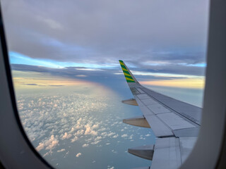 Wing view from an airplane window during flight, showcasing a serene sky with scattered clouds below and a soft rainbow glow on the horizon at golden hour.