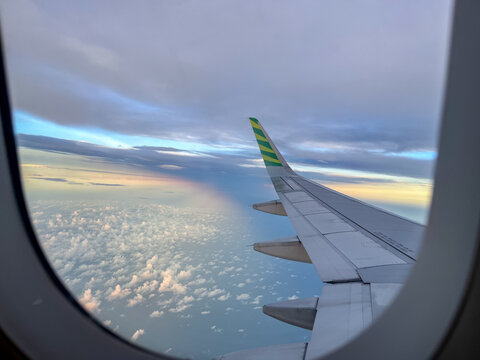 Wing view from an airplane window during flight, showcasing a serene sky with scattered clouds below and a soft rainbow glow on the horizon at golden hour.