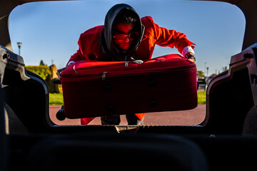 Naklejka premium Stylish latin tourist loading red suitcase into car trunk for road trip