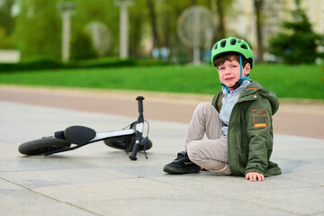 upset child with bicycle helmet sits on pavement after fall. young boy in green jacket, helmet beside fallen bike in park. childhood challenges, learning, outdoor activity theme.