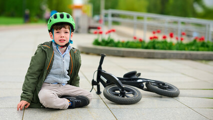 child with helmet sitting beside fallen balance bike on paved path outdoor. green park setting...
