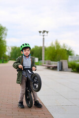Obraz premium boy with green helmet holding balance bike in park. child learning cycling skills, outdoor activity, healthy lifestyle concept. bright day, trees and benches in background