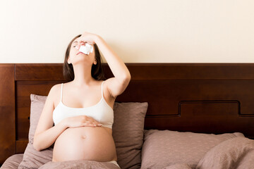 Portrait of young unhappy pregnant woman leaning on pillows on bed and blowing her nose into...