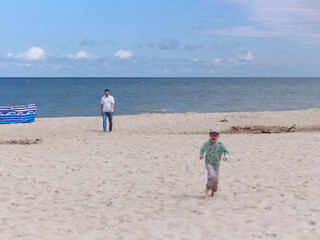 Father walking along a peaceful beach, following his joyful son who runs ahead out of focus, capturing a candid moment of family care and connection by the sea.