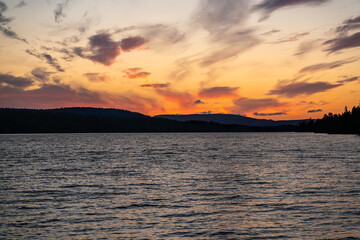 As the sun sets, vibrant colors reflect on Hällsjön lake, surrounded by the stunning Långfjället Nature Reserve in Sweden, offering a serene and picturesque view