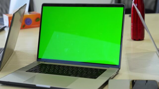 Laptop with green screen on an office desk with nearby tech items. Ideal for chroma key insertions, coworking scenes, startup environments, and modern digital workspace content.