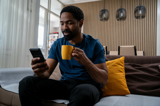 Smiling African American man sitting at home using smartphone and drinking coffee in living room.