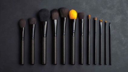 A collection of twelve black-handled makeup brushes of varying sizes and bristle types, arranged in a row on a dark gray surface; one brush head is a vibrant yellow-gold