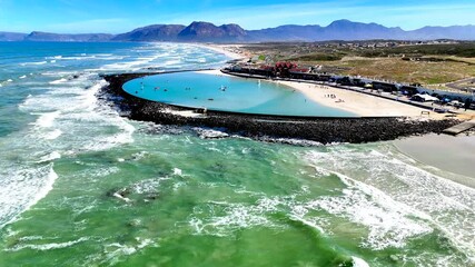 Drone view of the shoreline of a Cape Town beach with a large tidal pool and mountain range in the background on a summers day