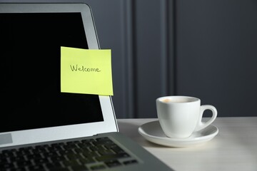 Sticky note with word Welcome, laptop and cup on white wooden table against grey background, closeup