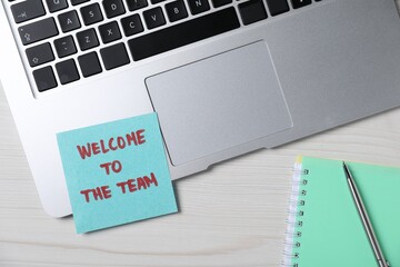 Paper note with words Welcome To The Team, laptop, notepad and pen on light wooden table, flat lay