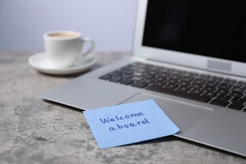 Sticky note with phrase Welcome Aboard, laptop and coffee on grey table, closeup