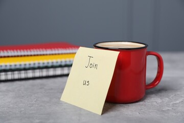 Sticky note with phrase Join Us, coffee and notebooks on grey table, closeup
