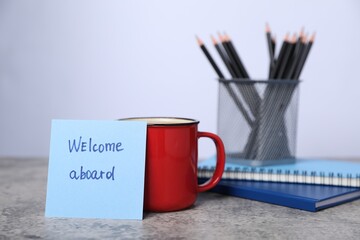 Sticky note with phrase Welcome Aboard, coffee, notebooks and pencils on grey table, closeup