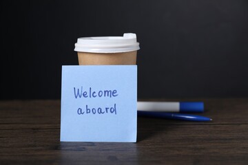 Paper note with phrase Welcome Aboard, cup of coffee and stationery on wooden table