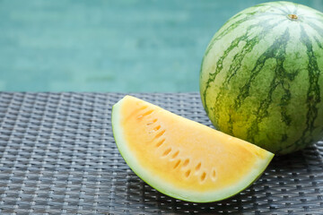 Whole and cut yellow watermelons on wicker table near swimming pool outdoors, closeup. Space for...