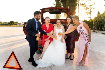 Bride and friends toasting with champagne by broken down car at sunset