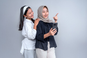 Two cheerful Asian women taking a selfie together, smiling warmly. One wears a hijab while...