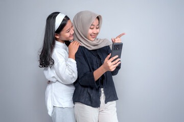 Two young Asian women smiling while taking a selfie. One wears a hijab and points at the phone screen. They appear happy, friendly, and close, standing in front of a plain background.