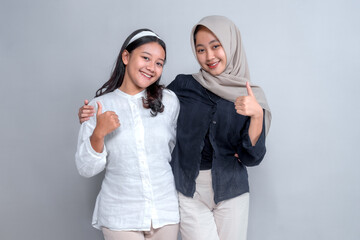 Two young Asian women smiling confidently while showing thumbs up. One wears a hijab, symbolizing...