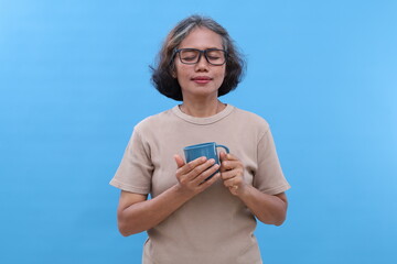 Asian elderly woman with grey hair, wearing glasses, smiling while holding drinking glass, closing...
