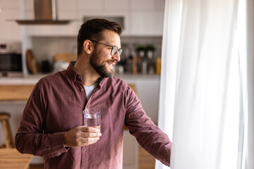 A bearded man wearing glasses drinks water from a glass in a modern kitchen. He enjoys a refreshing moment, focusing on hydration, wellness, and a healthy lifestyle in a bright, cozy home environment.